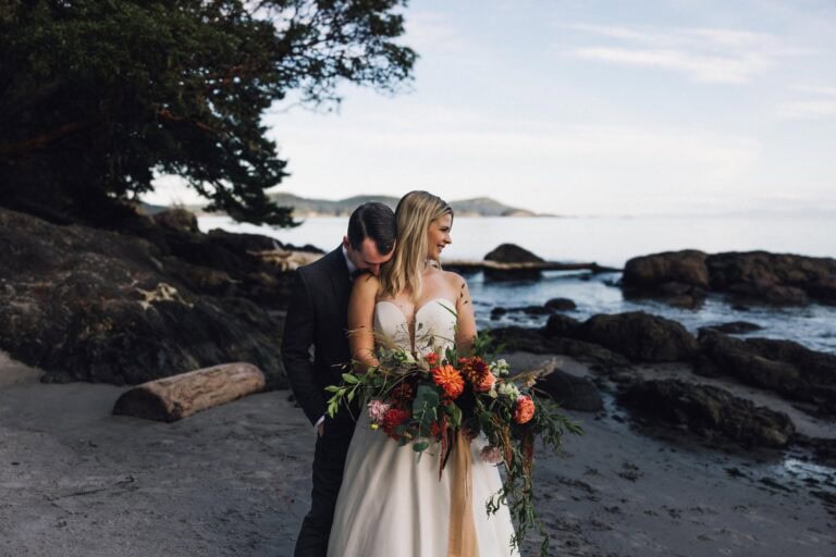 A couple standing near the ocean in sooke, bc for their elopement. He is hugging the bride from behind while she holds a large wedding bouquet.